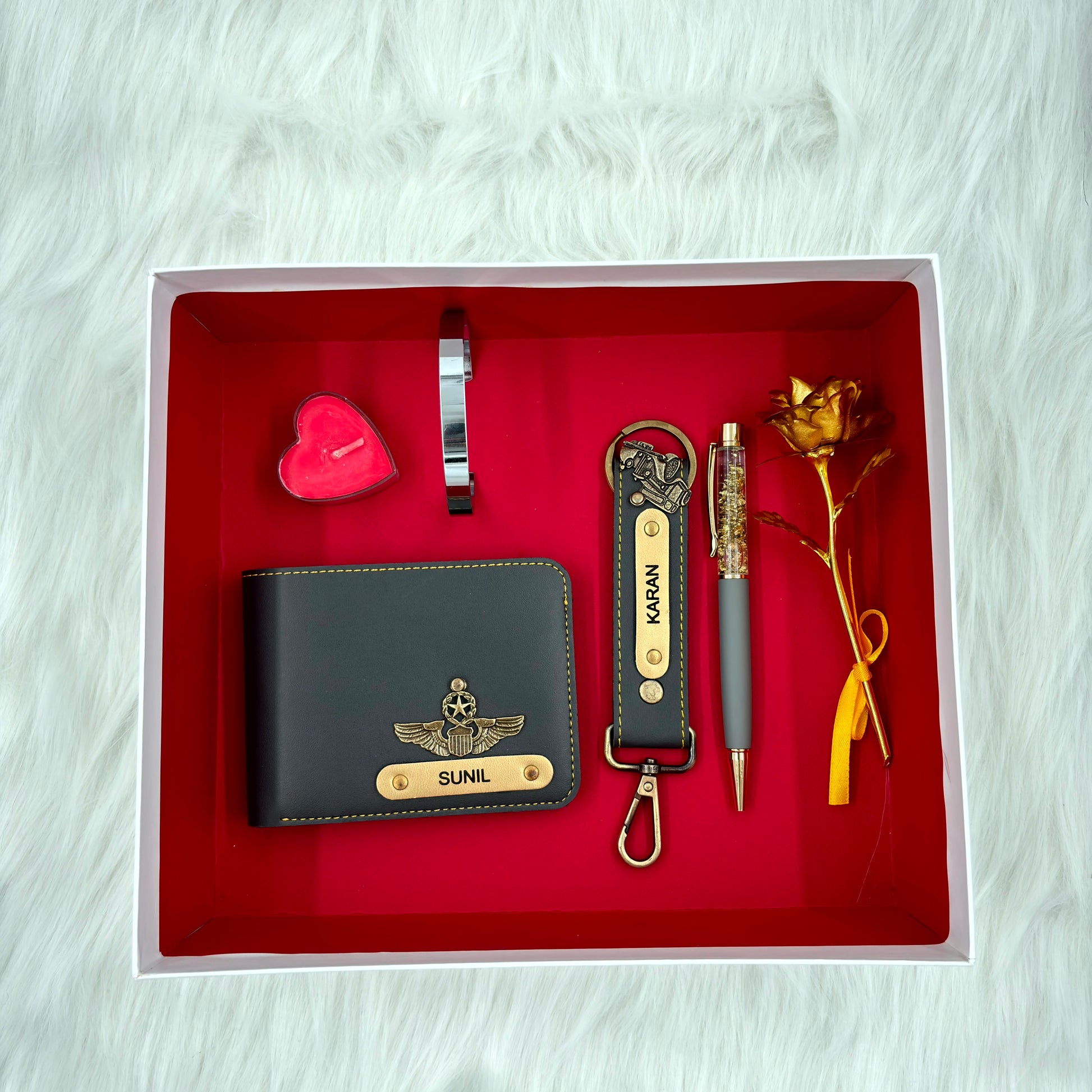 A top view of a box containing customized grey leather wallet, keychain, gold flake pen, kada, scented candle and gold plated rose.