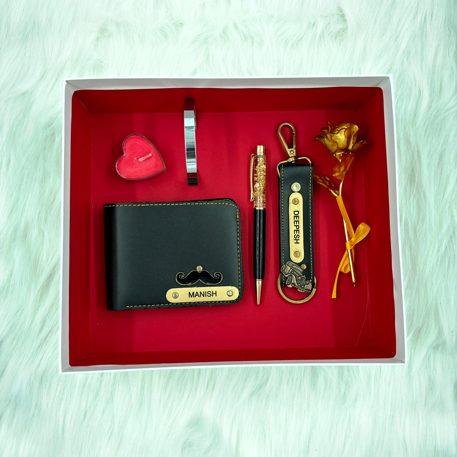 A top view of a box containing customized black leather wallet, keychain, gold flake pen, kada, scented candle and gold plated rose.
